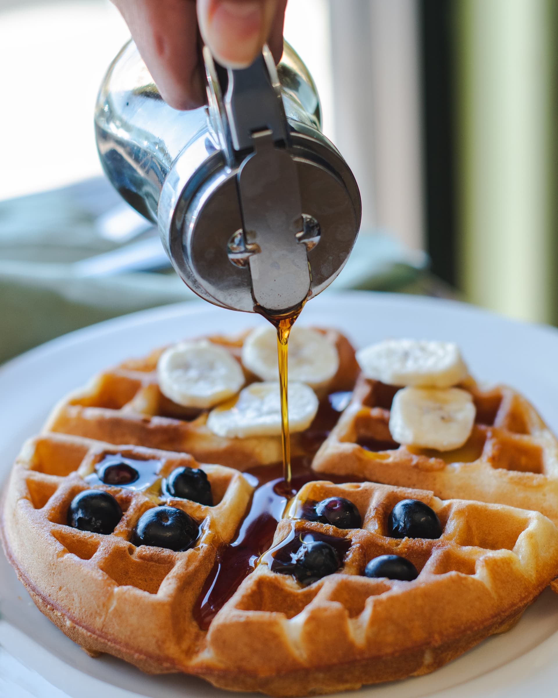 Vegan waffles topped with blueberries and bananas, syrup being poured